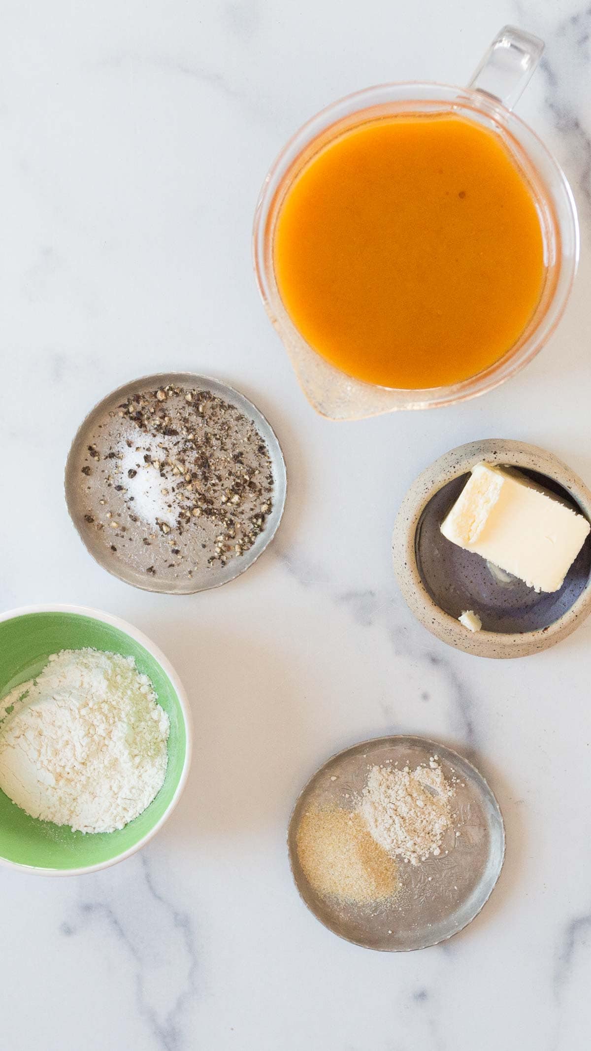 A top-down view of small bowls and a measuring cup containing ingredients for an Easy Gravy Recipe&mdash;ground pepper, salt, grated cheese, butter, an orange liquid, and a beige powder&mdash;on a white marble surface.