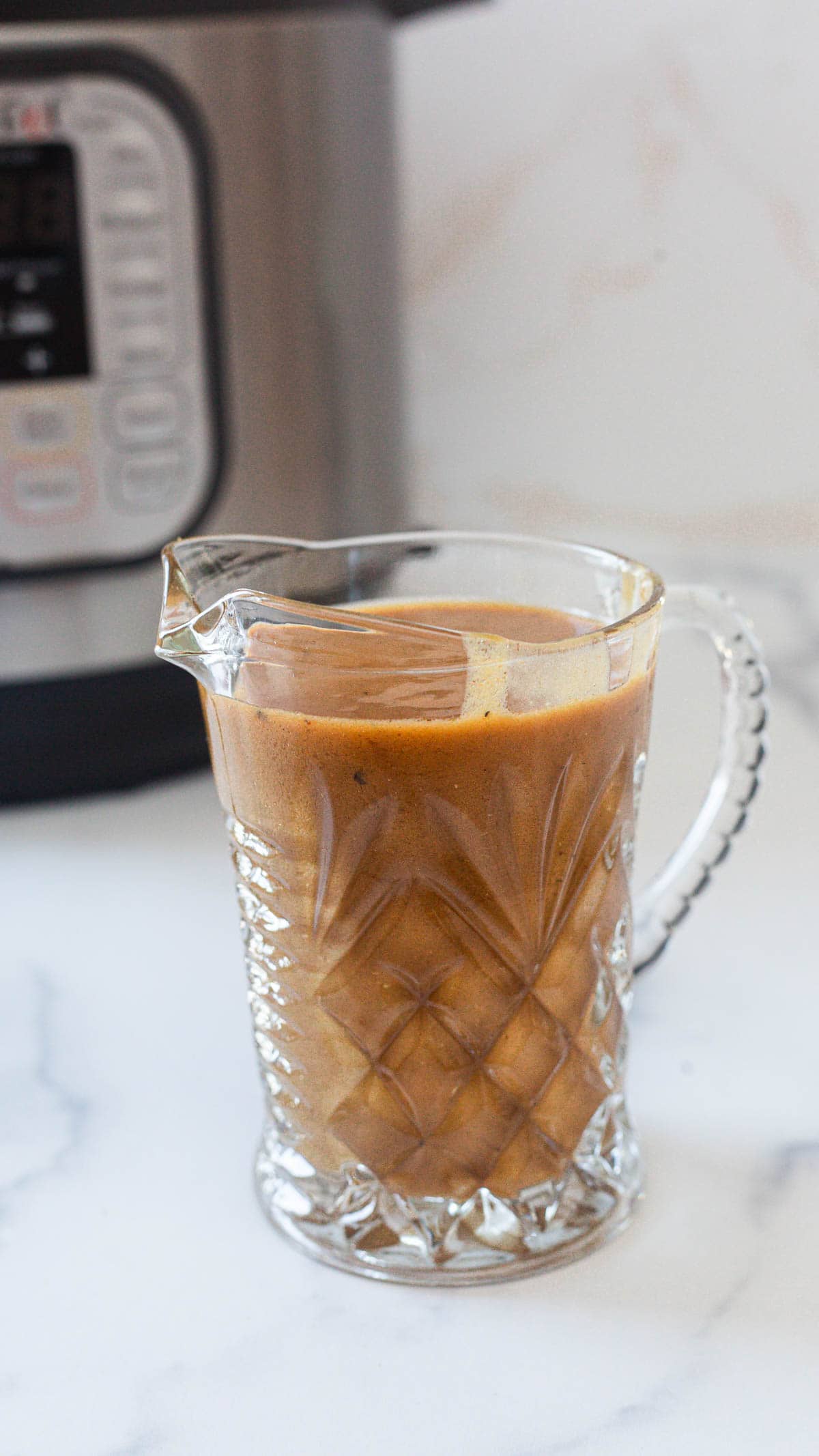 A glass pitcher filled with brown Instant Pot Gravy sits on a white countertop. An electric pressure cooker is visible in the background.