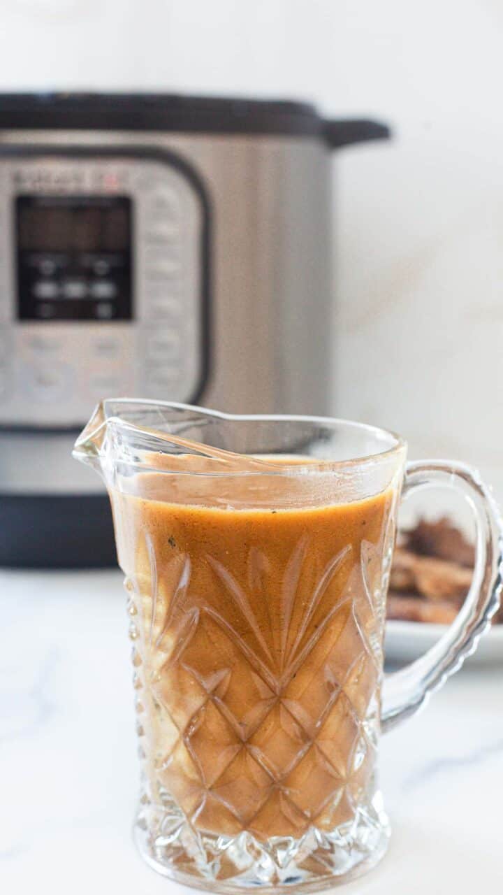 A glass pitcher filled with brown liquid sits on a white surface in front of an electric pressure cooker, hinting at a delicious Instant Pot Gravy. A small plate with spices is partially visible beside the pitcher.
