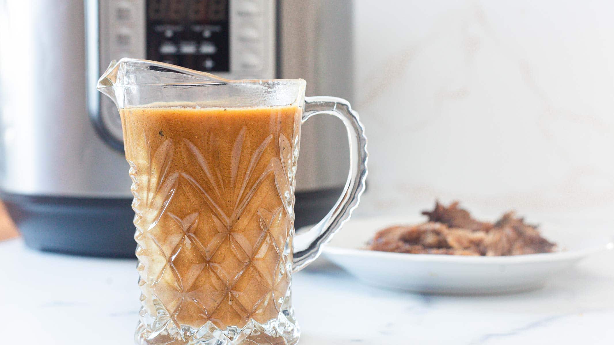 A glass pitcher filled with rich Instant Pot Gravy sits on a white countertop in front of a pressure cooker, with a plate of shredded meat blurred in the background&mdash;perfect for any easy gravy recipe.