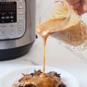 A hand pours brown Instant Pot Gravy from a glass container onto shredded meat on a white plate. An Instant Pot pressure cooker is visible in the background on a white surface.