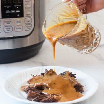 A hand pours creamy brown Instant Pot gravy from a glass pitcher onto shredded beef on a white plate, with an electric pressure cooker in the background, highlighting this easy gravy recipe.