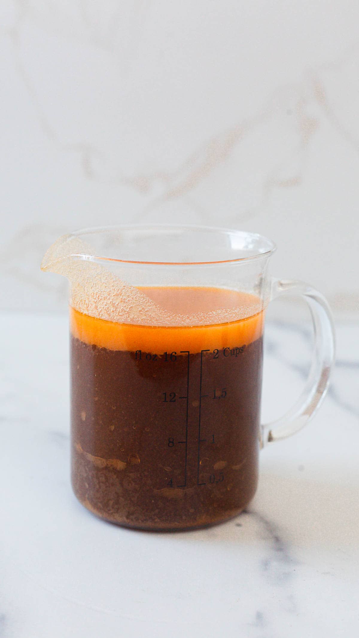 A glass measuring cup filled with dark brown Instant Pot gravy topped with a layer of orange oil, sitting on a white surface with a light marble background. The measurements are marked in cups and ounces&mdash;an easy gravy recipe visualized.
