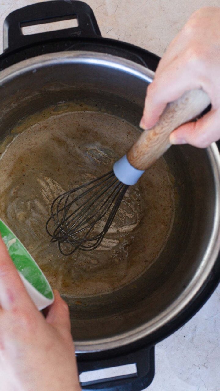 A person is making an Easy Gravy Recipe, whisking a brown liquid in a metal pot&mdash;possibly an Instant Pot&mdash;while pouring an ingredient from a green-and-white container into the mixture.