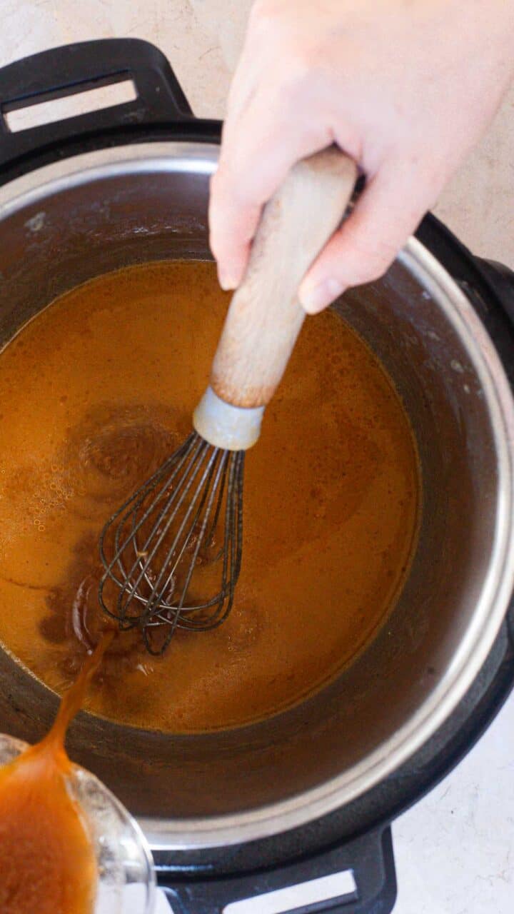 A hand holding a whisk stirs a brown liquid in an Instant Pot, while another hand pours more liquid from a glass measuring cup&mdash;demonstrating an easy gravy recipe perfect for making delicious Instant Pot gravy.