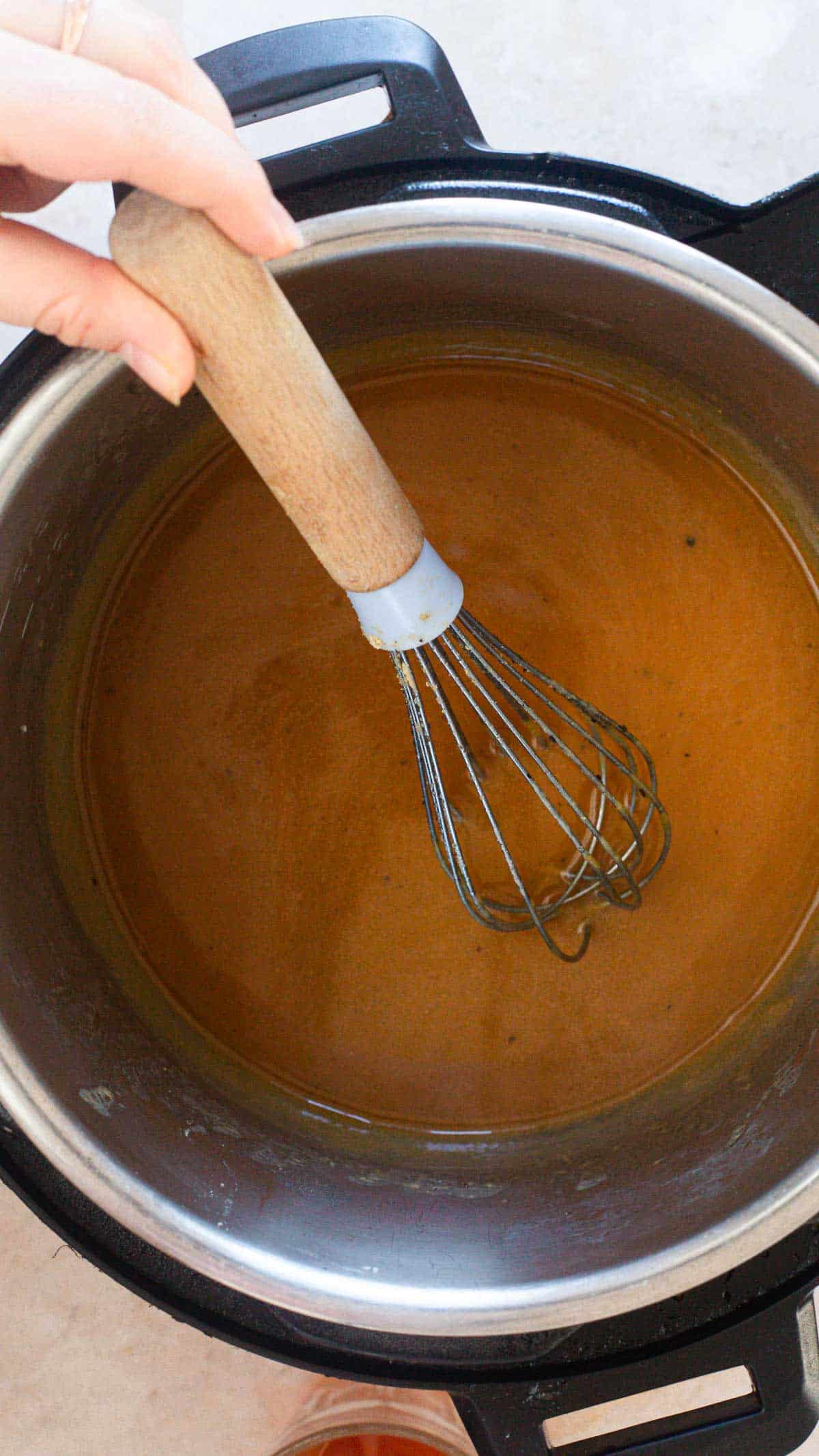A hand holding a whisk is stirring a pot of brown liquid inside an Instant Pot, making an Easy Gravy.