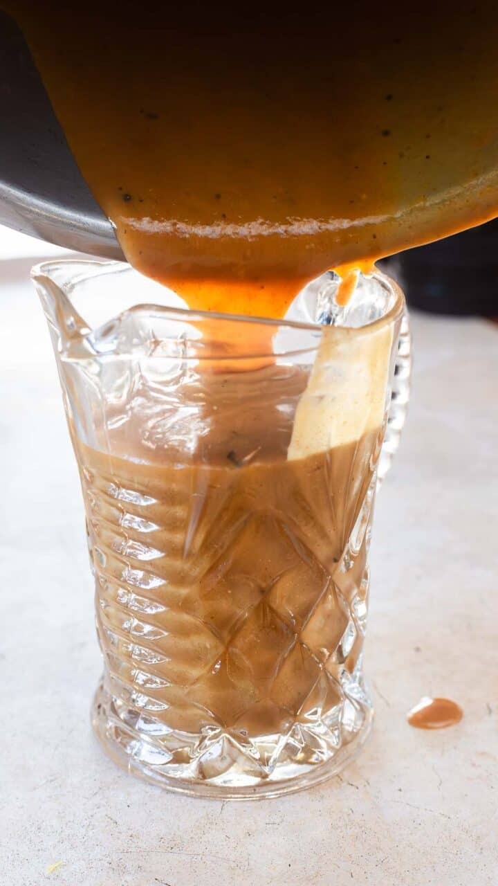 A thick brown gravy made from an Easy Gravy Recipe is being poured from a pan into a clear, diamond-patterned glass gravy boat on a light countertop. Some gravy is spilling over the side.