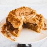 A round Biscoff Cake with light brown frosting and crumbled biscuit topping sits on a white cake stand. The cake has a slice missing, features a large Lotus Biscoff biscuit on top, and scattered crumbs decorate the platter.