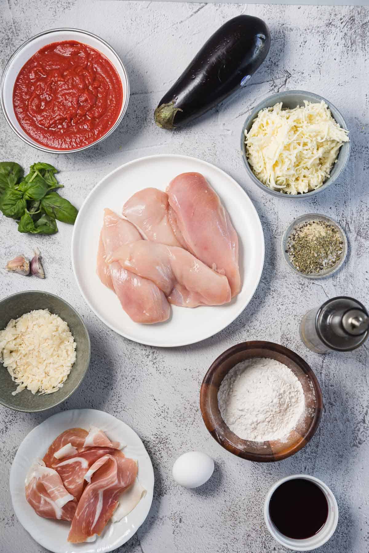 A flat lay of raw chicken breasts, mozzarella and parmesan cheese, marinara sauce, an eggplant, prosciutto, fresh basil, flour, an egg, minced garlic, dried herbs, black pepper, and a small bowl of red wine on a light surface.