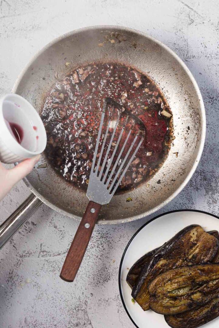 A hand pours liquid from a small white cup into a frying pan filled with a dark sauce and a spatula. Nearby, a plate holds several slices of cooked eggplant.