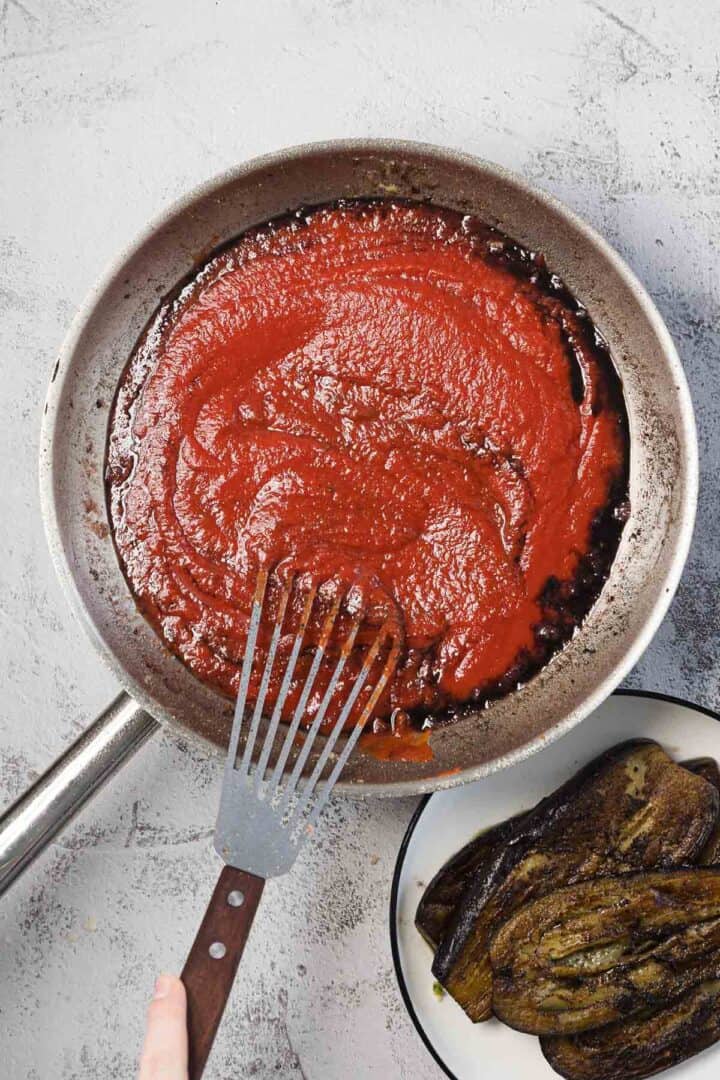 A hand holding a metal spatula stirs tomato sauce in a frying pan, with a plate of cooked eggplant slices next to the pan on a light gray countertop.