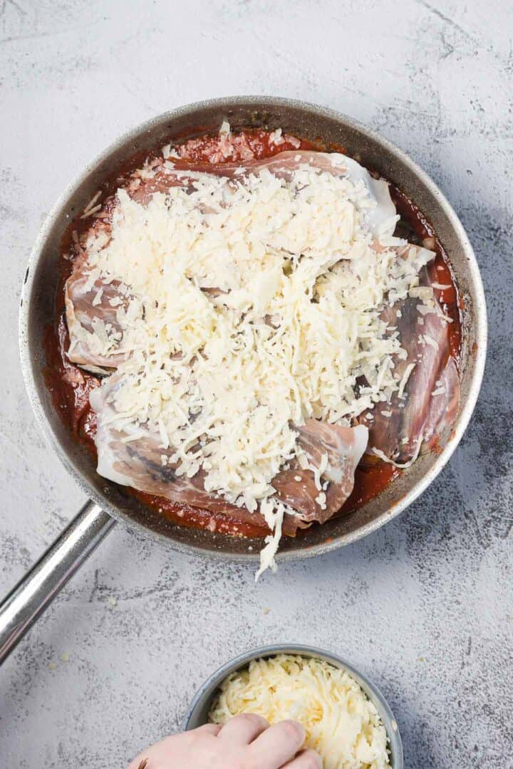 A metal skillet filled with tomato sauce, pieces of raw white fish, and topped with shredded cheese. A hand at the bottom adds more shredded cheese from a small bowl. The background surface is light grey.