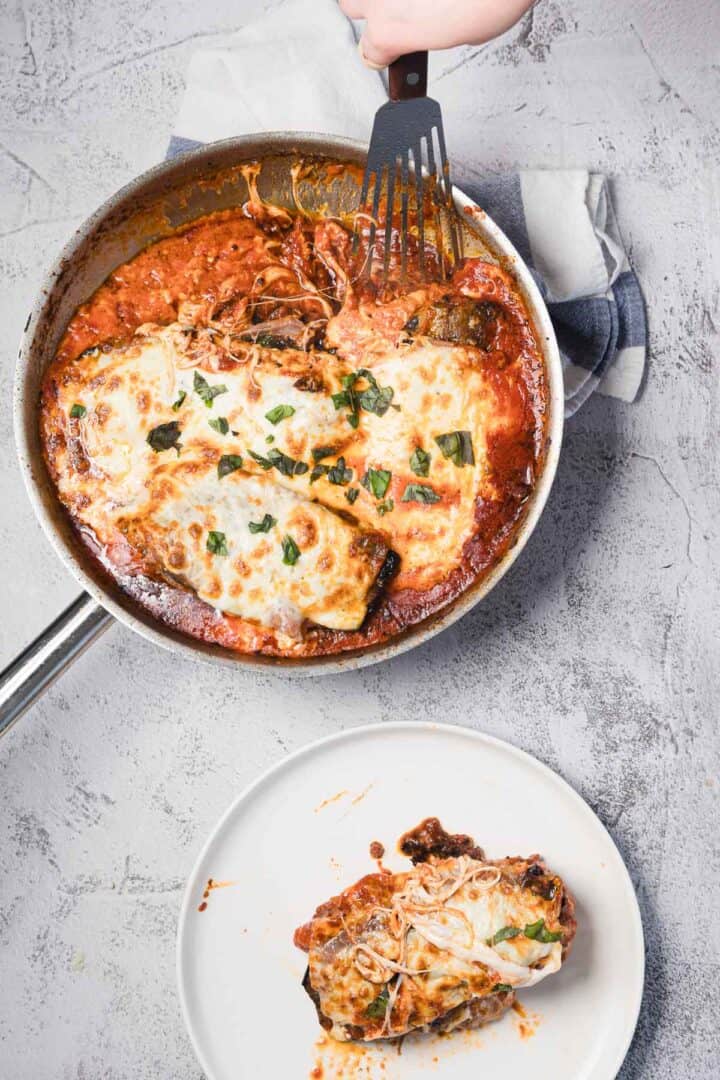 A skillet containing two portions of baked eggplant parmesan with melted cheese and tomato sauce, with one portion being served onto a white plate below. A hand holds a spatula above the skillet.