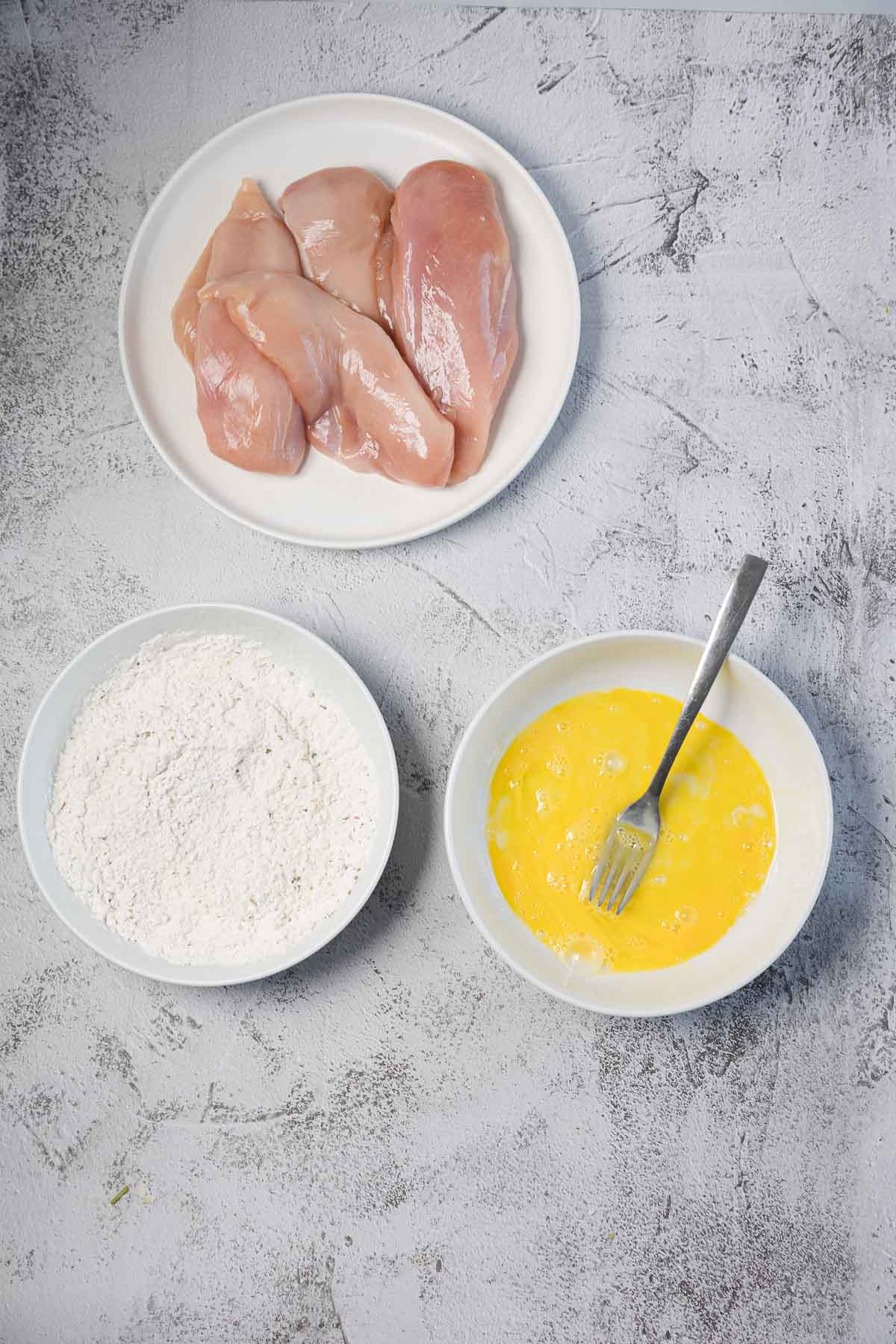 Three bowls are arranged on a gray surface: one with raw chicken breasts, one with flour, and one with beaten eggs and a fork.
