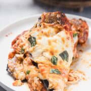 A serving of baked eggplant parmesan sits on a white plate, topped with melted cheese, tomato sauce, and fresh basil leaves, with spaghetti noodles partially visible underneath.