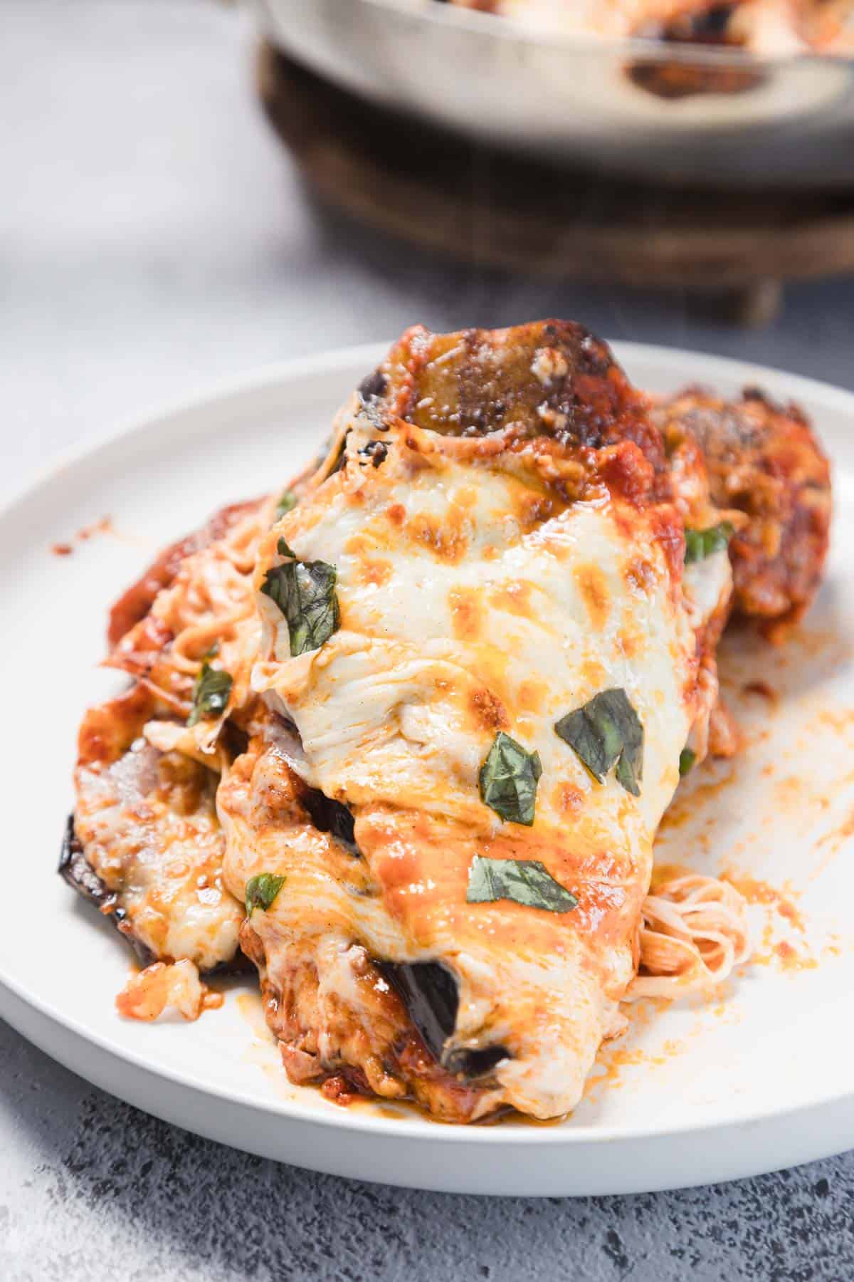 A serving of baked eggplant parmesan sits on a white plate, topped with melted cheese, tomato sauce, and fresh basil leaves, with spaghetti noodles partially visible underneath.