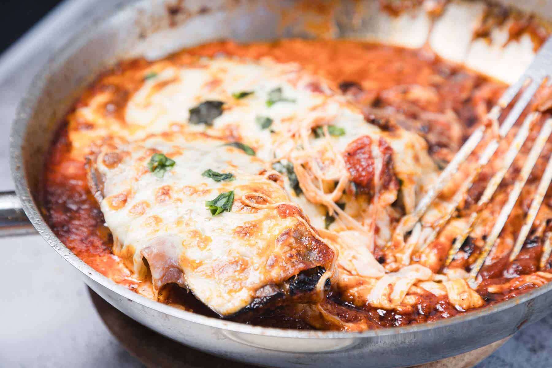 A metal pan filled with baked eggplant parmesan topped with melted cheese and tomato sauce. Some cheese is being lifted with a serving utensil, showing the cheesy texture and fresh basil garnish.