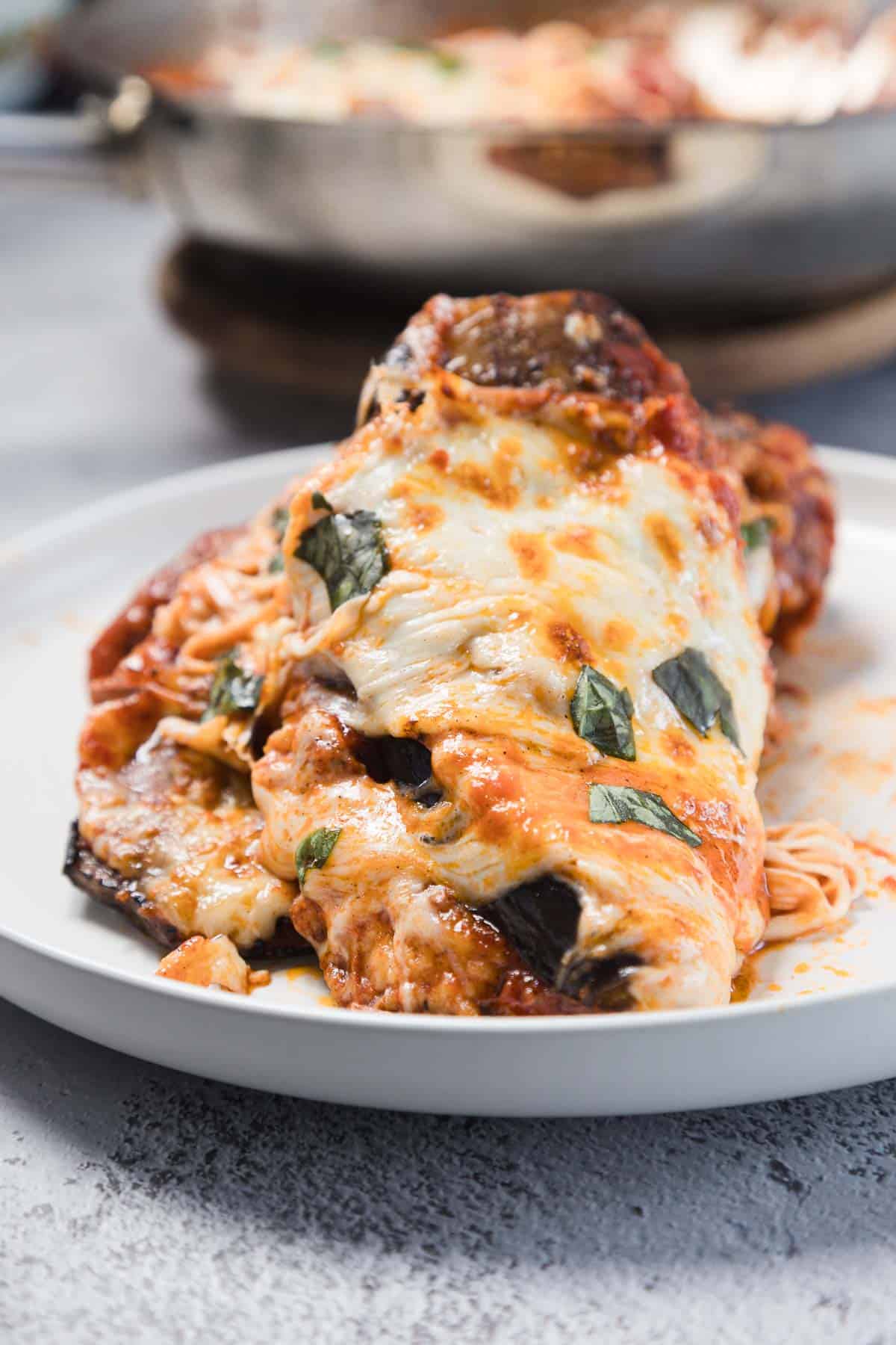 A close-up of eggplant parmesan topped with melted cheese, tomato sauce, and basil, served on a white plate with a portion of spaghetti visible underneath.