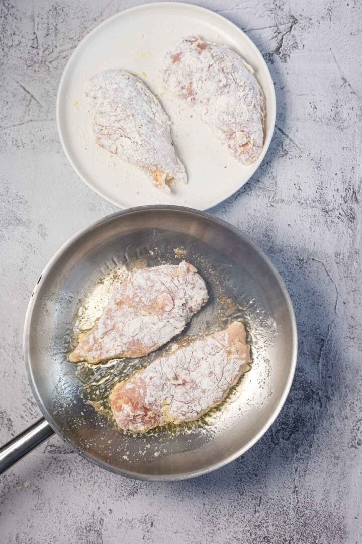 Two pieces of flour-coated raw chicken are in a frying pan with oil, while two more flour-coated chicken pieces rest on a white plate nearby, all set on a textured gray surface.