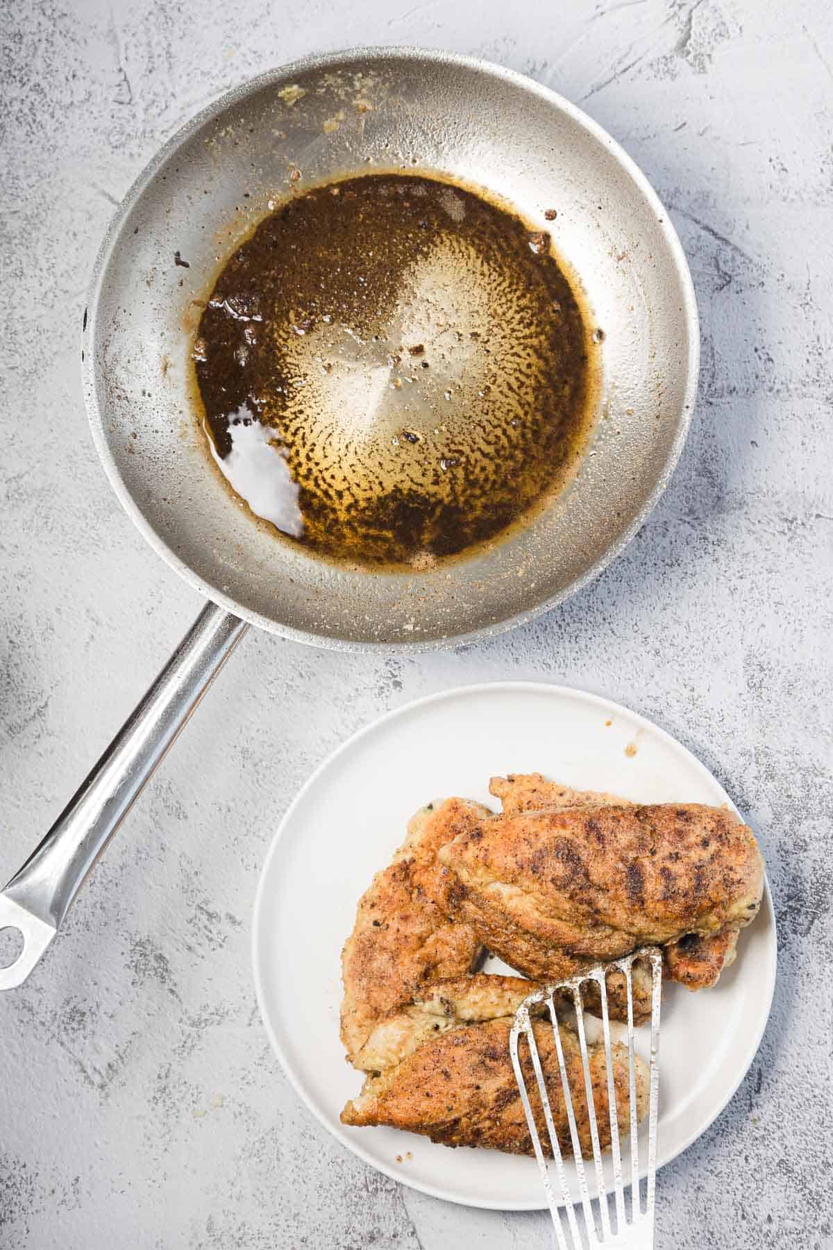 A metal frying pan with brown cooking residue sits above a white plate holding seasoned, cooked chicken breasts. A metal fork rests on one piece of chicken. The background surface is light gray.