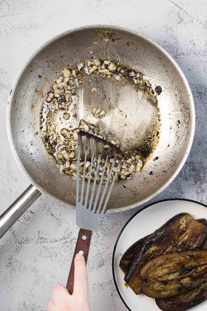 A hand holds a metal spatula in a stainless steel pan with saut&eacute;ed chopped garlic and oil. A bowl with cooked eggplant slices sits nearby on a light gray surface.