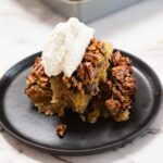 A serving of pecan pie bars, inspired by the classic Pecan Pie recipe, is topped with whipped cream on a black plate, with a baking dish of more bars in the background on a marble surface.