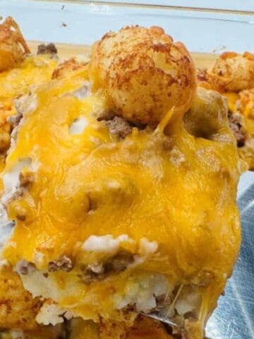 A close-up view of a casserole topped with melted cheddar cheese, tater tots, and ground beef being served from a glass baking dish with a metal spatula.
