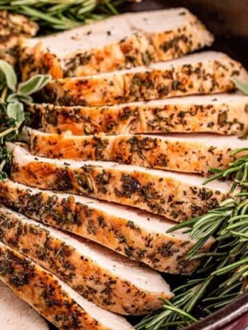 Slices of herb-crusted cooked turkey breast arranged in a cast iron skillet, garnished with fresh rosemary and thyme, with a ramekin of sauce visible in the background.