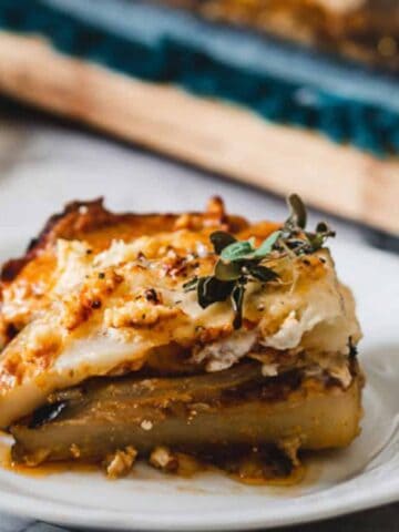 A slice of baked casserole with layers of vegetables, cheese, and sauce is served on a white plate. The dish is garnished with fresh herbs, and a cutting board with more casserole is in the background.