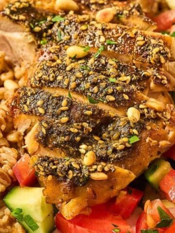 Sliced seasoned chicken breast served with a grain pilaf and a salad of diced tomatoes, cucumbers, and onions on a white plate with a fork.