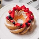 A Bundt cake topped with powdered sugar, sliced strawberries, raspberries, and blueberries sits on a white plate, with part of a patterned plate and fork visible in the background.