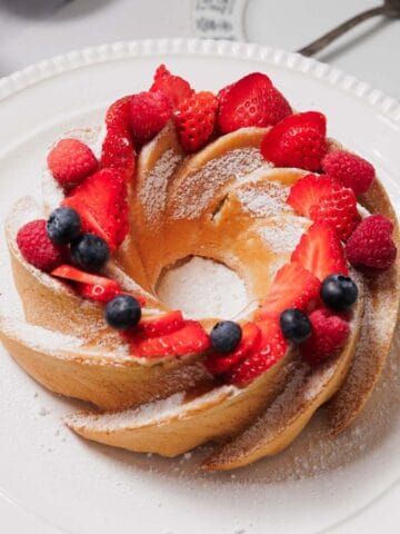 A Bundt cake topped with powdered sugar, sliced strawberries, raspberries, and blueberries sits on a white plate, with part of a patterned plate and fork visible in the background.