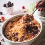 A hand holding a spoon takes a serving from a baked cherry cobbler in a white oval dish. Fresh cherries and a cup are visible in the softly focused background.