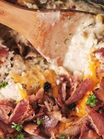 A close-up of a creamy dish topped with shredded cheddar cheese, chopped cooked bacon, and parsley, being stirred with a wooden spoon.