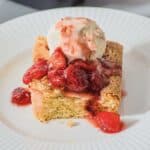 A slice of yellow sheet cake topped with a scoop of vanilla ice cream and strawberry sauce sits on a white plate, making it an easy twist on a classic Strawberry Sheet Cake.