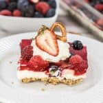 A berry pretzel dessert with a pretzel crust, cream cheese layer, red gelatin and berries, topped with whipped cream and a halved strawberry, served on a white plate. A bowl of mixed berry salad is in the background.