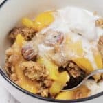 A bowl of peach cobbler, a classic peach dessert, features visible peach slices, crumbly topping, pecans, and a scoop of melting vanilla ice cream on top, with a spoon in the bowl.