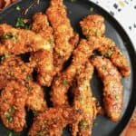 A black plate filled with several pieces of crispy, golden-brown Air Fryer Chicken Tenders, garnished with chopped parsley. The plate is partially on a white and black patterned surface.