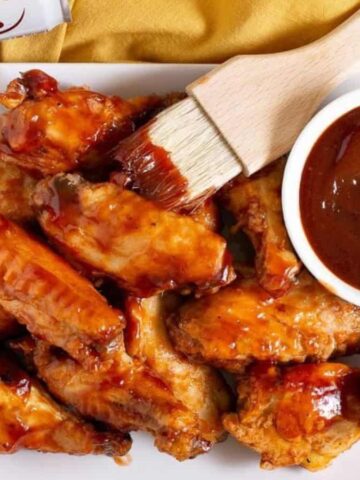 A square white plate with barbecue chicken wings, a small bowl of barbecue sauce, and a brush coated in sauce resting on top. A can is partially visible in the upper left corner on a yellow surface.
