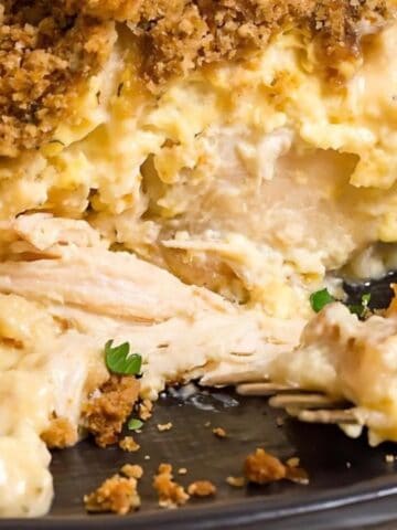 A close-up of a serving of creamy chicken casserole with a golden brown crumb topping on a dark plate, partially eaten with a fork lying beside it. Small green herb pieces are sprinkled on top.