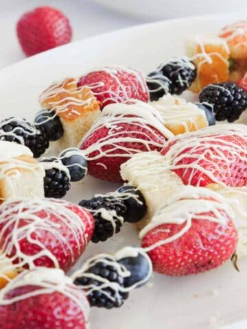 Skewers with strawberries, blackberries, blueberries, and cubes of cake are arranged on a white plate and drizzled with white chocolate. A bowl of blackberries and a single strawberry are in the background.