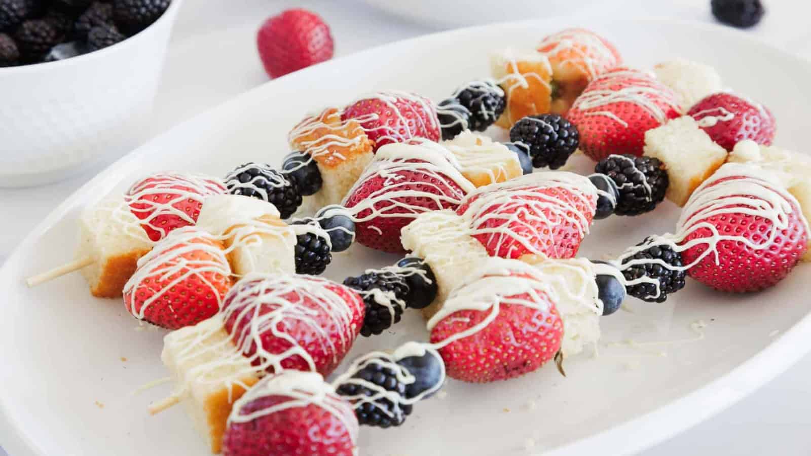 Skewers with strawberries, blackberries, blueberries, and cubes of cake are arranged on a white plate and drizzled with white chocolate. A bowl of blackberries and a single strawberry are in the background.