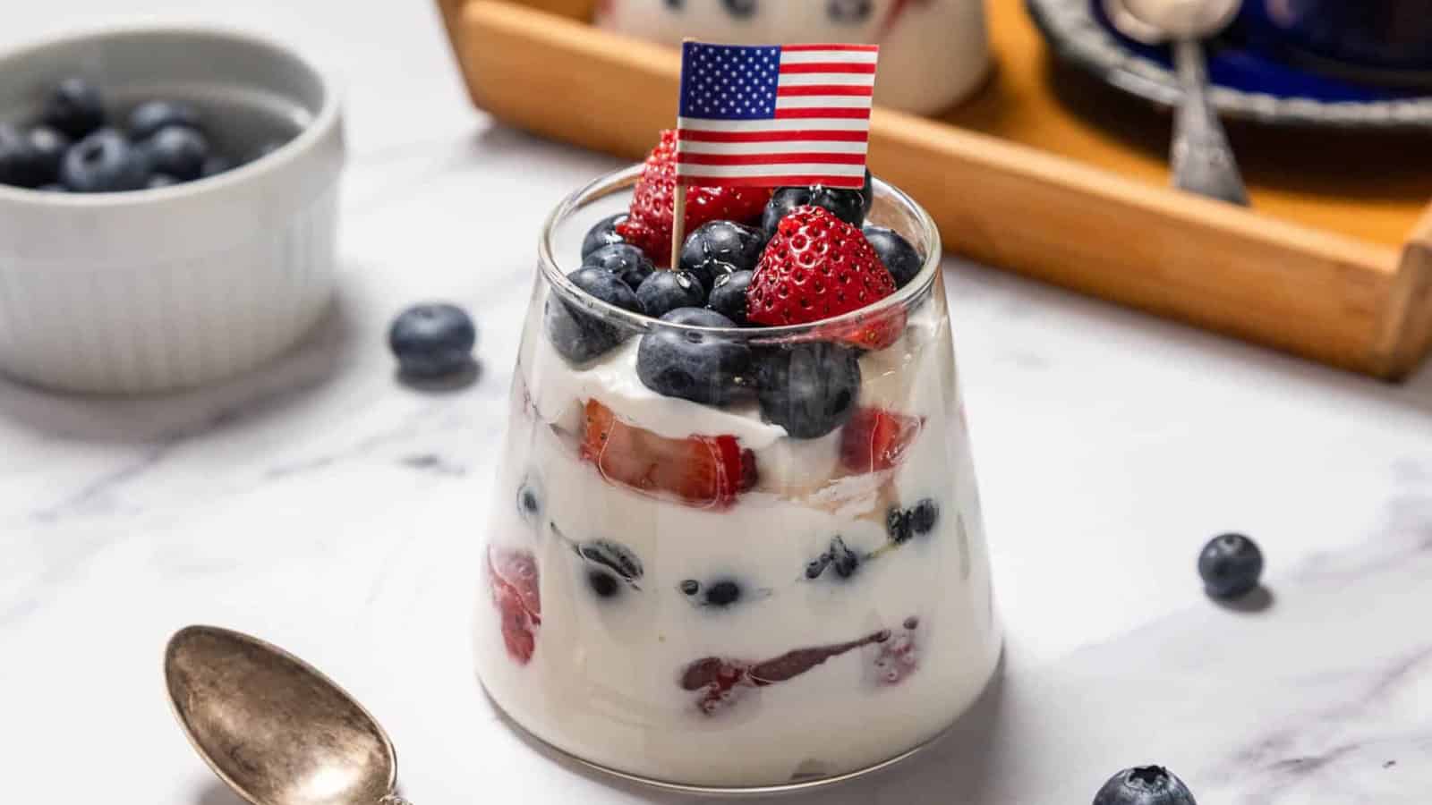 A glass filled with layers of yogurt, strawberries, and blueberries, topped with more berries and a small American flag on a toothpick. A spoon and a bowl of blueberries are nearby on a marble surface.