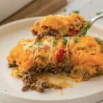 A fork holds a portion of this casserole recipe, featuring sweet potato, ground beef, melted cheese, red peppers, and chopped parsley, above a plate with the remaining serving of the dish.