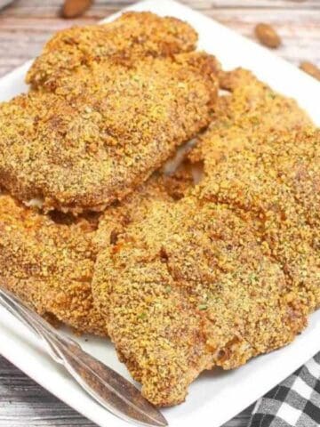 A white plate holds four pieces of breaded and baked chicken. A fork is placed beside the chicken, and whole almonds are scattered on the wooden table next to a black and white checkered cloth.