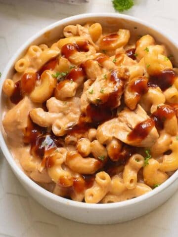 A bowl of macaroni and cheese with pieces of chicken, topped with a drizzle of brown sauce, sits on a white surface beside a fork and sprigs of parsley.