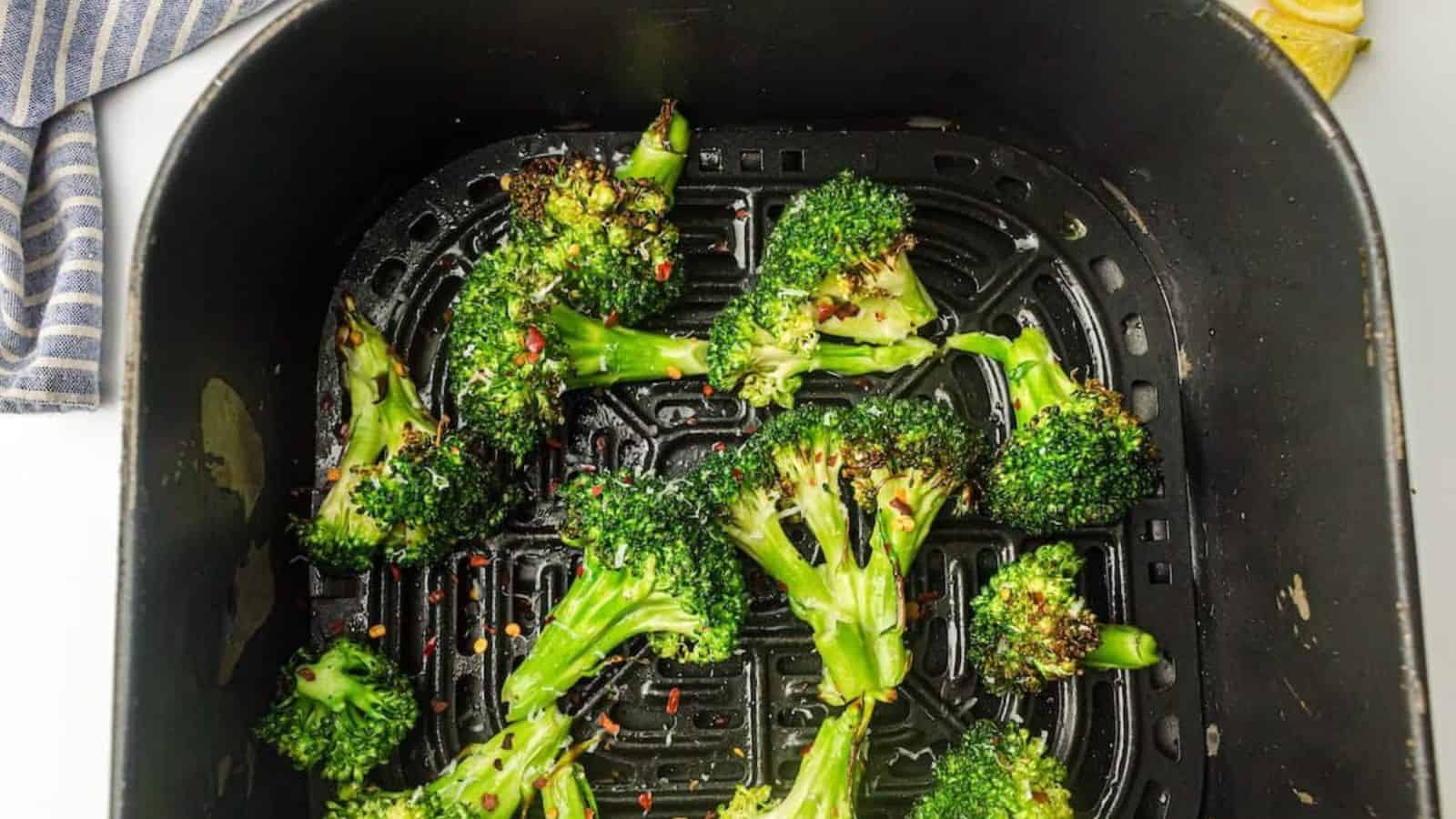 Broccoli florets with seasoning in an air fryer basket, some lemon slices and a striped cloth nearby.