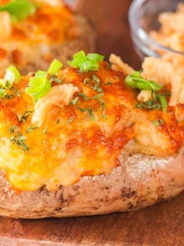 A baked potato topped with melted cheese, green onions, and crispy fried onions sits on a wooden board. Another stuffed potato is partially visible, with a glass bowl of fried onions in the background.