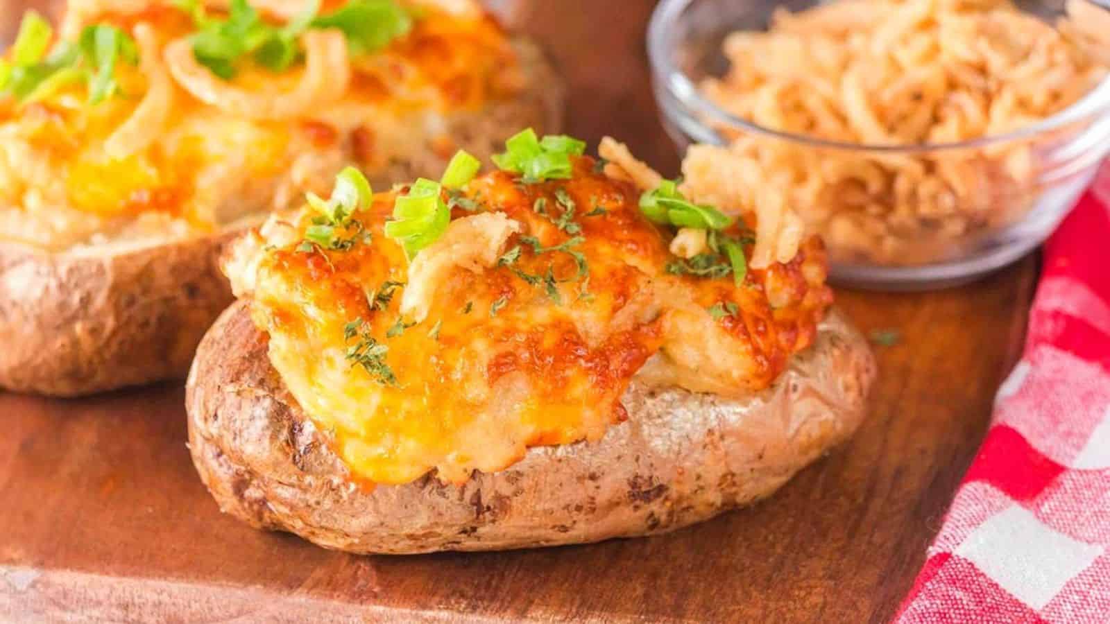 Loaded baked potatoes topped with melted cheese, green onions, and crispy fried onions, served on a wooden surface with a small bowl of more crispy onions in the background.