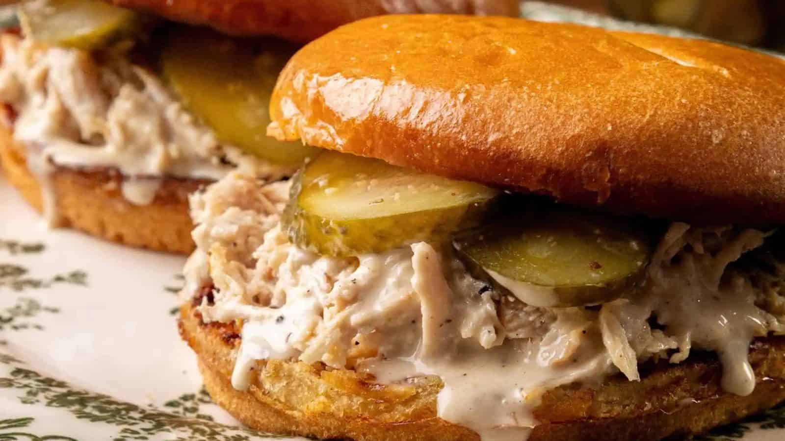 Two Alabama Chopped Chicken Sandwiches with pulled chicken, creamy Alabama white BBQ sauce, and pickle slices on shiny, toasted buns, served on a decorative plate.
